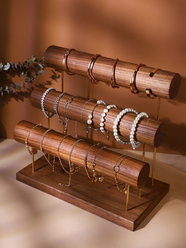 May include: A three-tiered jewellery display stand made of dark wood with gold-coloured metal supports. The stand holds various bracelets, including gold bangles, beaded bracelets, and pearl strands. The wooden rods are cylindrical.