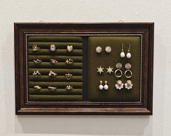 May include: A display case with a dark brown wooden frame, showcasing various gold-toned rings and earrings. The rings are arranged on a green velvet surface, while the earrings are displayed on a similar green background. The jewelry includes studs, hoops, and drop earrings.