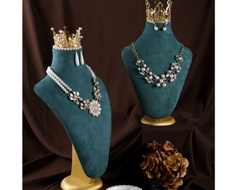 May include: Two teal jewelry busts display necklaces, earrings, and crowns. One bust features a double-strand pearl necklace with a floral pendant. The other bust showcases a pearl and gold necklace. A small dish holds a pearl necklace.