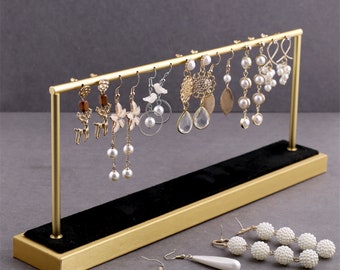 May include: Gold metal earring stand with a black velvet base. The stand has a horizontal bar with 9 pairs of earrings hanging from it. The earrings are a variety of styles and colors, including pearl, crystal, and floral designs.