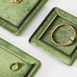 May include: Gold jewelry set on a green velvet display. The set includes a necklace with a rectangular green stone, a bracelet with clear stones, and a ring. The jewelry is displayed on square trays.