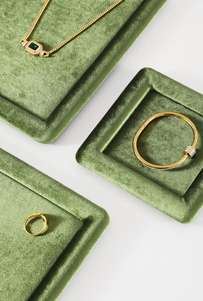 May include: Gold jewelry set on a green velvet display. The set includes a necklace with a rectangular green stone, a bracelet with clear stones, and a ring. The jewelry is displayed on square trays.