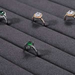 Puede incluir: Cuatro anillos con piedras preciosas de diferentes colores expuestos en un soporte de anillos de terciopelo gris. Los anillos son de plata y oro con piedras preciosas transparentes y verdes.