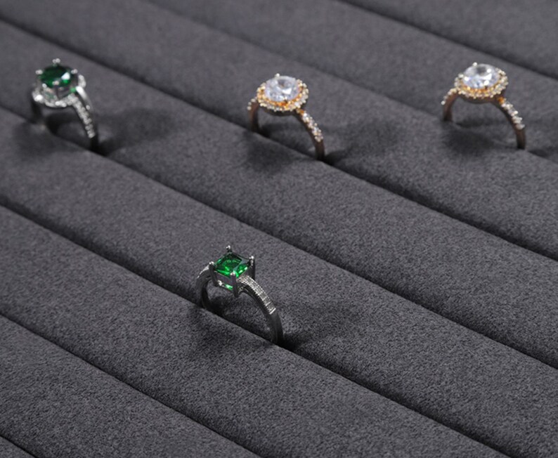 Puede incluir: Cuatro anillos con piedras preciosas de diferentes colores expuestos en un soporte de anillos de terciopelo gris. Los anillos son de plata y oro con piedras preciosas transparentes y verdes.