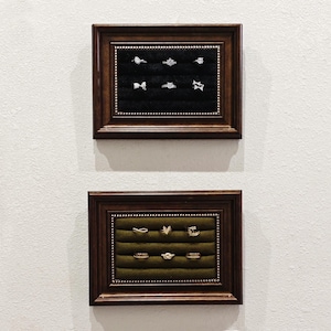 May include: Two dark wood frames with velvet lining display a variety of rings. The top frame has a black velvet lining and displays silver rings. The bottom frame has a green velvet lining and displays gold rings.