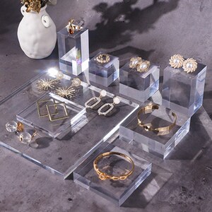 May include: A set of clear acrylic display stands for jewelry. The stands are of various sizes and shapes, and are arranged on a gray surface. The stands are displaying a variety of jewelry, including earrings, rings, and bracelets.
