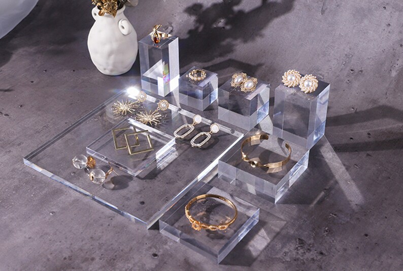 May include: A set of clear acrylic display stands for jewelry. The stands are of various sizes and shapes, and are arranged on a gray surface. The stands are displaying a variety of jewelry, including earrings, rings, and bracelets.