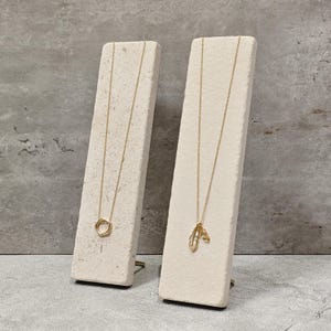 Könnte beinhalten: Zwei goldene Halsketten, präsentiert auf cremefarbenen, rechteckigen Ständern. Eine Halskette hat einen kreisförmigen Anhänger, die andere ein blattartiges Design. Die Ständer sind abgewinkelt und zeigen den Schmuck vor einem strukturierten, grauen Hintergrund.