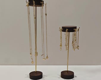 Op de afbeelding: Twee sieradenstandaards. De standaards hebben een donkerbruine houten basis en bovenkant met een goudkleurige metalen paal. De standaards bevatten meerdere gouden kettingen van verschillende lengtes en ontwerpen. De achtergrond is effen lichtgrijs.