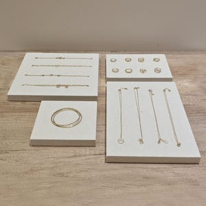 Puede incluir: Una muestra de joyas de oro en soportes blancos. Las joyas incluyen collares, pulseras y anillos. Las joyas están dispuestas en una cuadrícula.