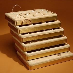 Op de afbeelding: Een stapel van zes beige fluwelen juwelendoosjes met gouden randjes. Elk doosje heeft meerdere vakjes om ringen, oorbellen en armbanden op te bergen.