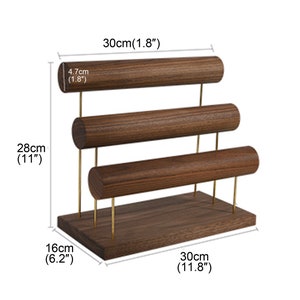 May include: A wooden jewellery display stand with three horizontal bars for bracelets or bangles. The stand is 28cm tall, 30cm wide, and 16cm deep. The bars are 4.7cm in diameter. The stand has gold-coloured metal supports.