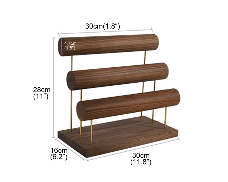 May include: A wooden jewellery display stand with three horizontal bars for bracelets or bangles. The stand is 28cm tall, 30cm wide, and 16cm deep. The bars are 4.7cm in diameter. The stand has gold-coloured metal supports.