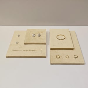 Peut inclure: Divers bijoux présentés sur des plateformes rectangulaires beiges. Les bijoux comprennent des boucles d'oreilles, un bracelet et des bagues. Les boucles d'oreilles présentent des motifs floraux et ronds. Le bracelet est délicat et les bagues sont des anneaux simples.