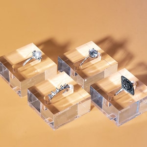 Puede incluir: Cuatro anillos exhibidos sobre bloques de acrílico transparente. Los anillos presentan varios diseños, incluyendo una perla, una gema y detalles metálicos ornamentados. Los anillos son plateados y están sobre un fondo cálido, mostrando joyería.