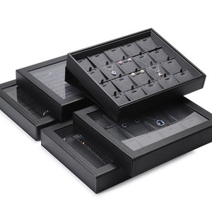 Puede incluir: Joyero de cuero negro de tres niveles. El nivel superior tiene una tapa transparente y est&aacute; dividido en compartimentos para collares y pendientes. El nivel intermedio tiene una tapa transparente y est&aacute; dividido en compartimentos para anillos y pulseras. El nivel inferior tiene una tapa negra y es un compartimento grande abierto.