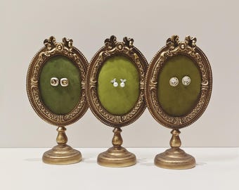 May include: Three gold-colored, ornate, oval-shaped display stands with green velvet interiors. Each stand has a pair of earrings displayed on the velvet.