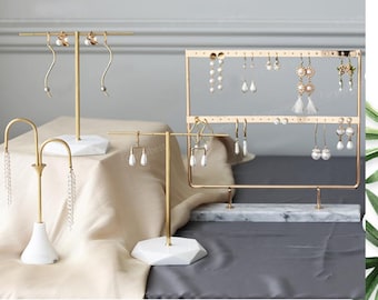 May include: Three gold metal earring stands with white marble bases. The stands are designed to hold earrings of different styles. The stand on the left has a curved design, the stand in the center has a straight bar, and the stand on the right has a rectangular frame with multiple hooks.