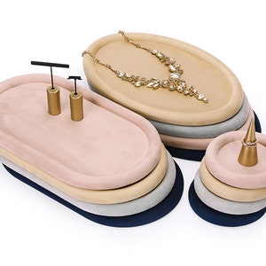 May include: A set of four oval-shaped jewelry trays in navy, gray, beige, and pink with a gold ring holder and earring stands. The trays are stacked on top of each other.