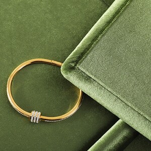 May include: A gold-colored bracelet with a row of small, clear stones and three vertical bars of clear stones. The bracelet is set against a green velvet background.