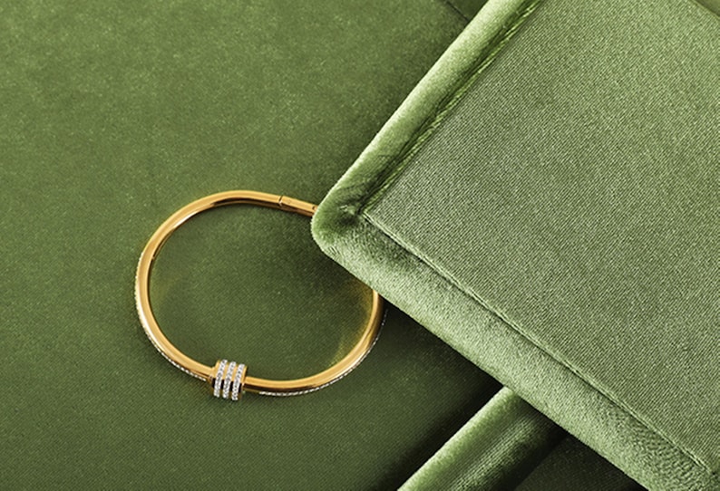 May include: A gold-colored bracelet with a row of small, clear stones and three vertical bars of clear stones. The bracelet is set against a green velvet background.