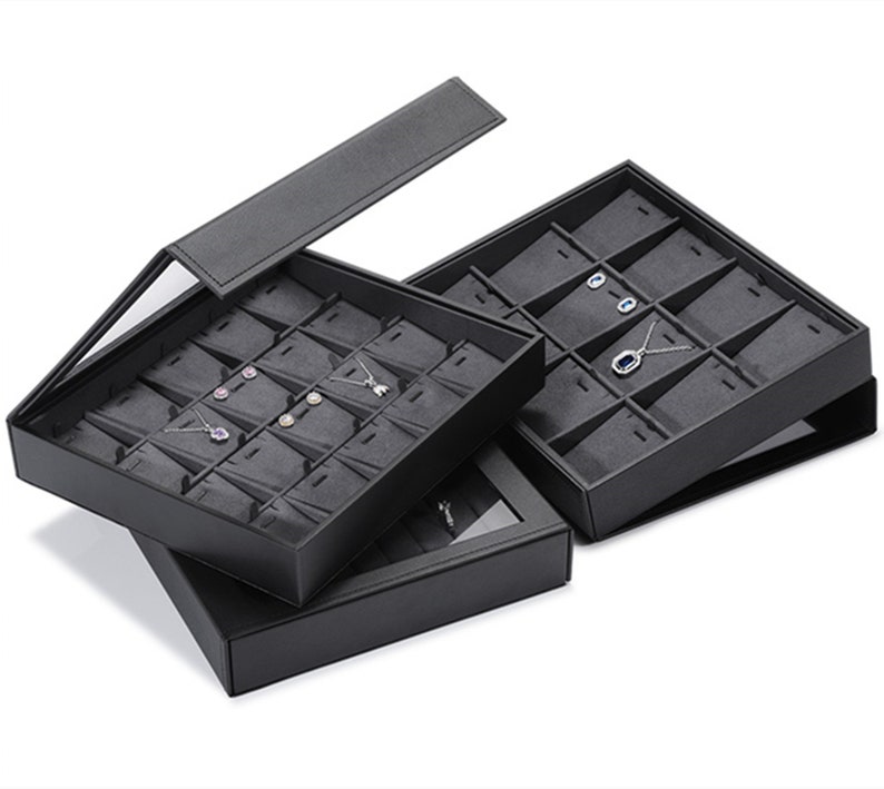 Puede incluir: Caja de almacenamiento de joyas negra con tapa transparente. La caja tiene varios compartimentos para guardar pendientes, collares y otras joyas.