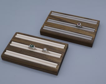 May include: Two wooden ring display trays with beige velvet lining. Each tray has four slots for rings. One tray has three rings, one with a green stone, one with a white stone, and one with a red stone. The other tray has one ring with a red stone.