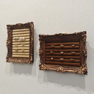 May include: Two ornate, gold-framed ring displays mounted on a wall. One has a beige velvet interior, the other a brown velvet interior. Each display holds multiple rings and earrings, showcasing jewellery in an elegant manner.