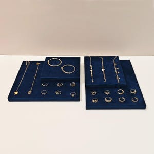 Może przedstawiać: Niebieski, welurowy stojak na biżuterię z dwoma poziomami. Na górnym poziomie znajdują się dwie złote bransoletki, a na dolnym poziomie znajduje się wiele złotych pierścionków. Drugi stojak ma trzy złote naszyjniki, a na dolnym poziomie znajduje się wiele złotych pierścionków.