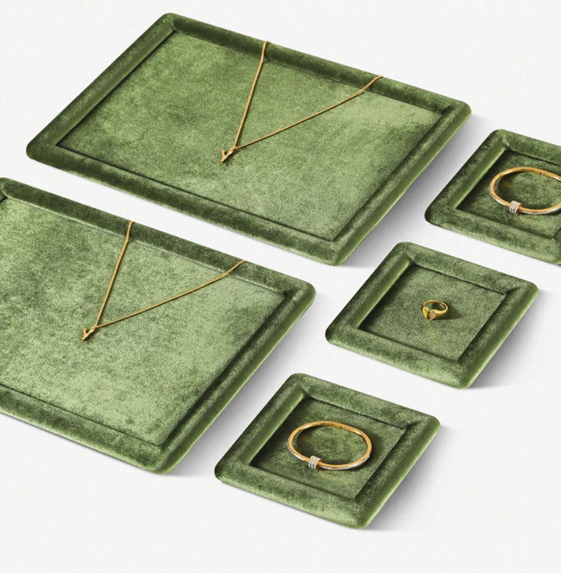 May include: An assortment of jewelry displayed on olive green velvet trays. The jewelry includes gold necklaces, bracelets, and a ring. The trays are rectangular and square, creating an organized presentation. The jewelry is set against a white background.