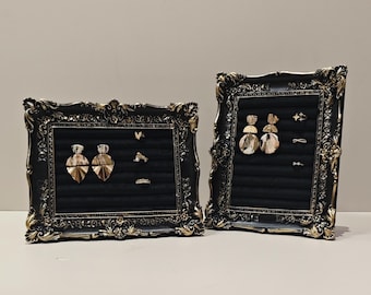 May include: Two ornate black and gold framed jewelry displays. Each frame holds a black velvet insert with various earrings and rings. The earrings are gold-toned and feature geometric and abstract designs. The frames have detailed, vintage-style embellishments.