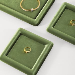 May include: Olive green velvet jewelry display trays with gold rings and a bracelet. The trays are square and rectangular, with a raised border. The jewelry is gold-toned, with a ring featuring a small gemstone.