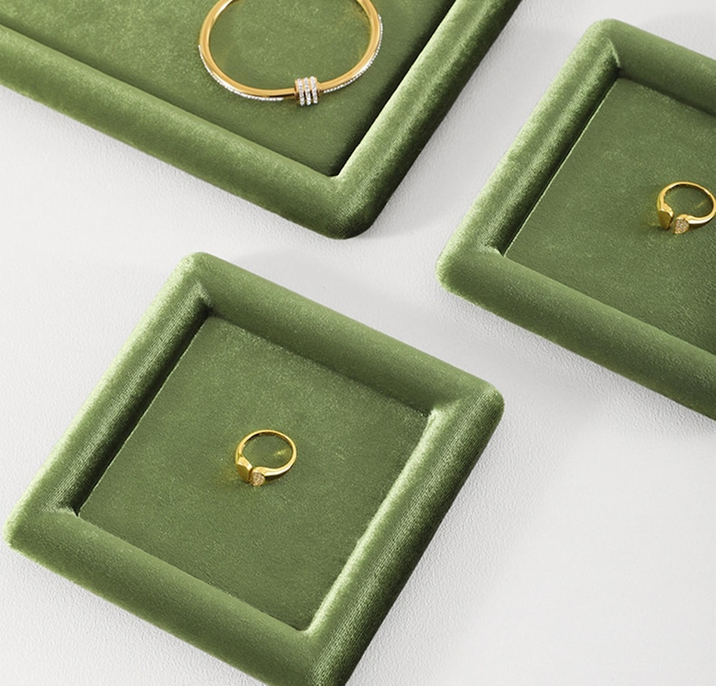 May include: Olive green velvet jewelry display trays with gold rings and a bracelet. The trays are square and rectangular, with a raised border. The jewelry is gold-toned, with a ring featuring a small gemstone.