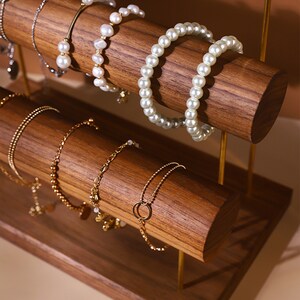 May include: A wooden jewellery display stand with multiple levels. The stand holds various bracelets, including pearl and gold chain styles. The display has a warm wood tone and gold metal supports, showcasing the jewellery.