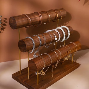 May include: A three-tiered wooden jewellery display stand with gold-coloured metal supports. The stand holds various bracelets and bangles in gold, silver, and pearl. The wood has a rich, dark brown finish.