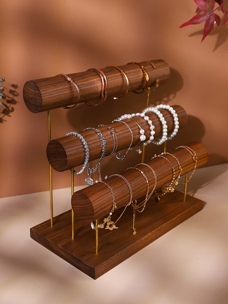 May include: A three-tiered wooden jewellery display stand with gold-coloured metal supports. The stand holds various bracelets and bangles in gold, silver, and pearl. The wood has a rich, dark brown finish.