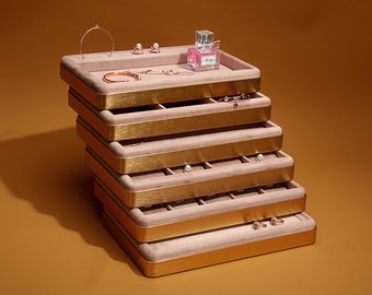 May include: A stack of five rectangular jewelry trays with brushed gold metal frames and soft pink velvet interiors. The trays feature various compartments for organizing accessories, with a small perfume bottle and several pieces of jewelry placed on top.