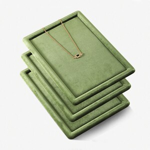 May include: Three stacked, rectangular, olive-green velvet jewelry display trays. A delicate gold necklace with a small, dark gemstone pendant rests on the top tray. The trays are angled, showcasing the necklace and the texture of the velvet.