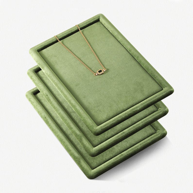 May include: Three stacked, rectangular, olive-green velvet jewelry display trays. A delicate gold necklace with a small, dark gemstone pendant rests on the top tray. The trays are angled, showcasing the necklace and the texture of the velvet.