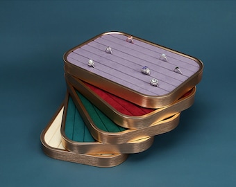 May include: A stack of four ring display trays with velvet lining. The top tray is lavender with gold trim and features six slots for rings. The other trays are red, green, and cream.