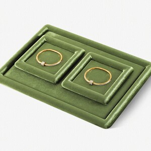 May include: Olive green velvet jewelry display with two gold bracelets. The display has a tiered design with a rectangular base and two square inserts. Each bracelet has a gold band with a small, sparkling accent.