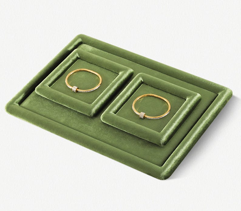 May include: Olive green velvet jewelry display with two gold bracelets. The display has a tiered design with a rectangular base and two square inserts. Each bracelet has a gold band with a small, sparkling accent.