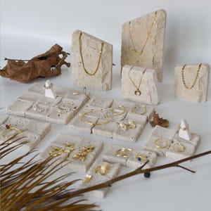 May include: A display of gold jewelry, including necklaces, rings, and bracelets, presented on textured, light-colored stone blocks. The jewelry features various designs, including chains, pendants, and pearl accents. A dried leaf and branch add a natural touch.