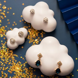 Op de afbeelding: Twee witte, wolkvormige oorbellenstandaards met verschillende oorbellen. De ene standaard heeft twee paar zilveren oorbelstuds met heldere stenen. De andere standaard heeft twee paar gouden oorhangers met blauwe stenen.