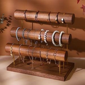 May include: A three-tiered wooden jewellery display stand with gold-coloured metal supports. The stand holds various bracelets and necklaces, including pearl and chain styles. The wood has a rich, dark brown colour.