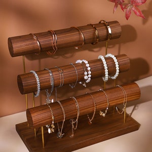 May include: A three-tiered jewellery display stand made of dark wood with gold-coloured supports. The stand holds various bracelets and necklaces. The bracelets are gold, silver, and pearl. The background is a warm brown.