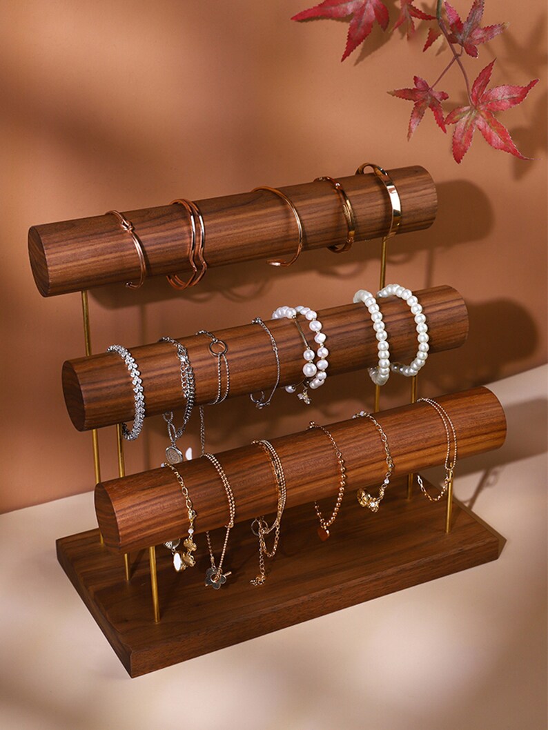 May include: A three-tiered jewellery display stand made of dark wood with gold-coloured supports. The stand holds various bracelets and necklaces. The bracelets are gold, silver, and pearl. The background is a warm brown.
