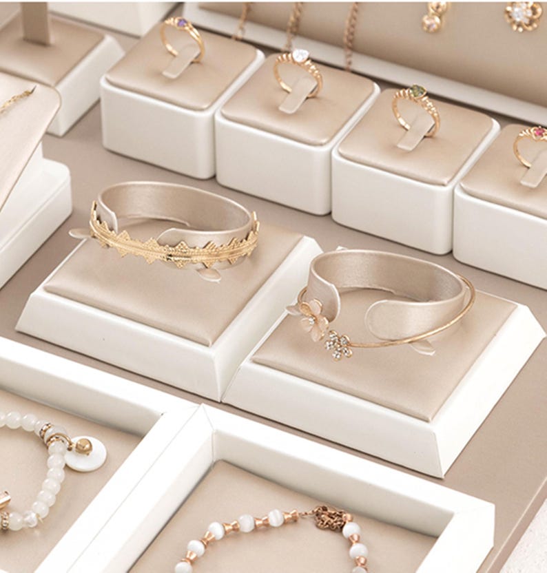 May include: White jewelry display with multiple compartments for rings, bracelets, and necklaces. The display has a beige velvet lining and is made of a sturdy material.
