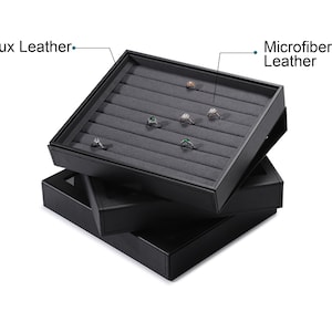 Puede incluir: Bandeja de exhibici&oacute;n de anillos de cuero sint&eacute;tico negro con forro de cuero de microfibra. La bandeja tiene m&uacute;ltiples ranuras para anillos y est&aacute; apilada sobre otras dos bandejas similares.