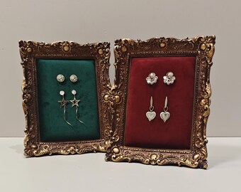 May include: Two ornate gold picture frames with velvet backing, one green and one red, displaying various earrings. The earrings include star-shaped, round, and heart-shaped designs.